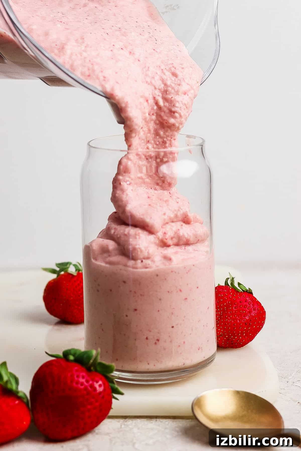 A thick pink strawberry smoothie is being poured from a blender into a glass, with fresh strawberries and a spoon beside the glass.