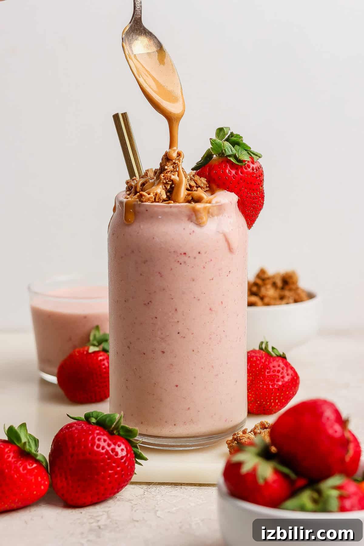 A glass of strawberry smoothie topped with granola and peanut butter, surrounded by fresh strawberries, with a spoon drizzling peanut butter on top.