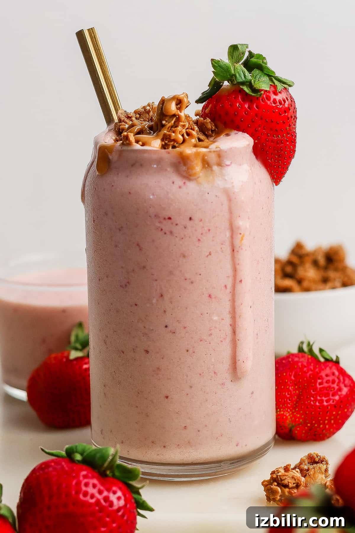A glass of strawberry smoothie with granola, peanut butter drizzle, a gold straw, and a whole strawberry on top, surrounded by fresh strawberries and a bowl of granola.