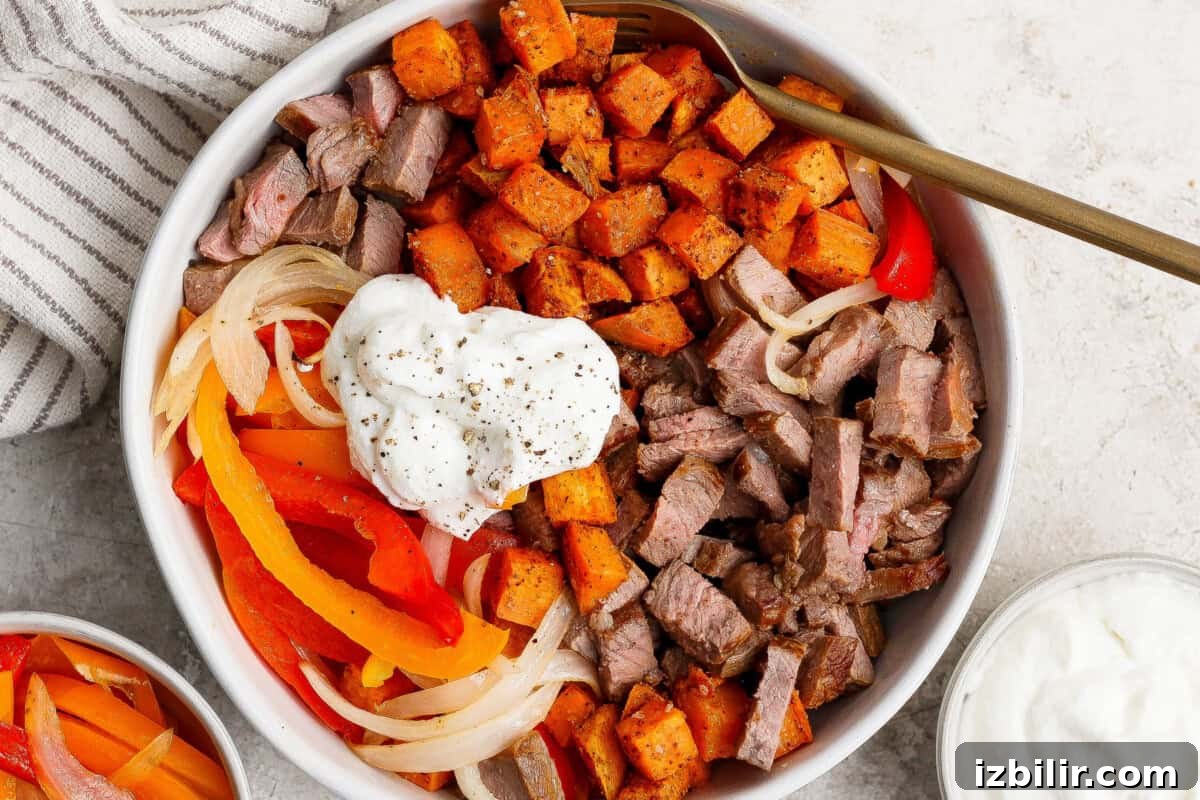 A bowl filled with diced steak, roasted sweet potatoes, sautéed onions and bell peppers, topped with a dollop of sour cream and black pepper, with a fork resting inside.