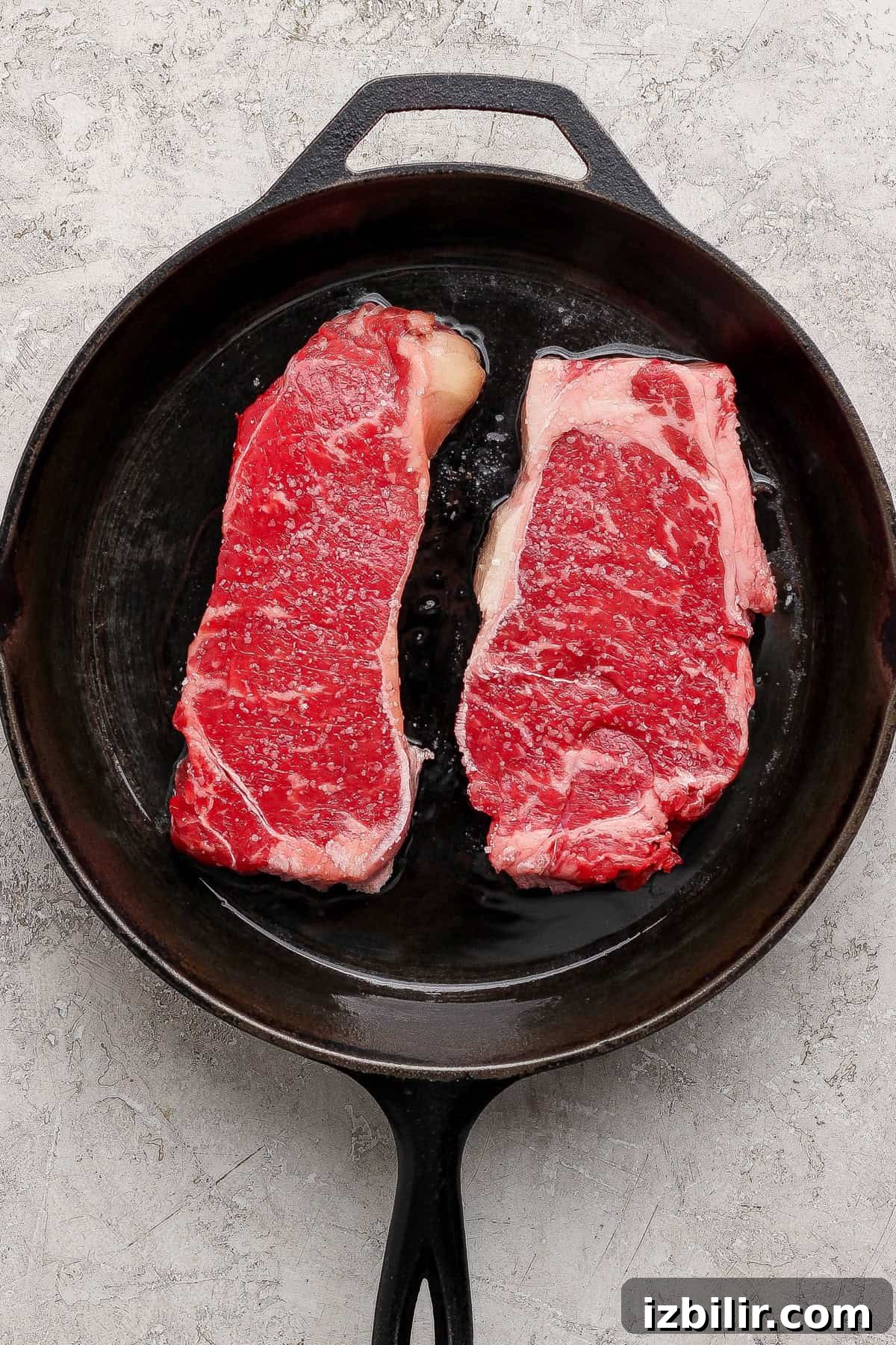 Two raw steaks are placed side by side in a black cast iron skillet on a light textured surface.