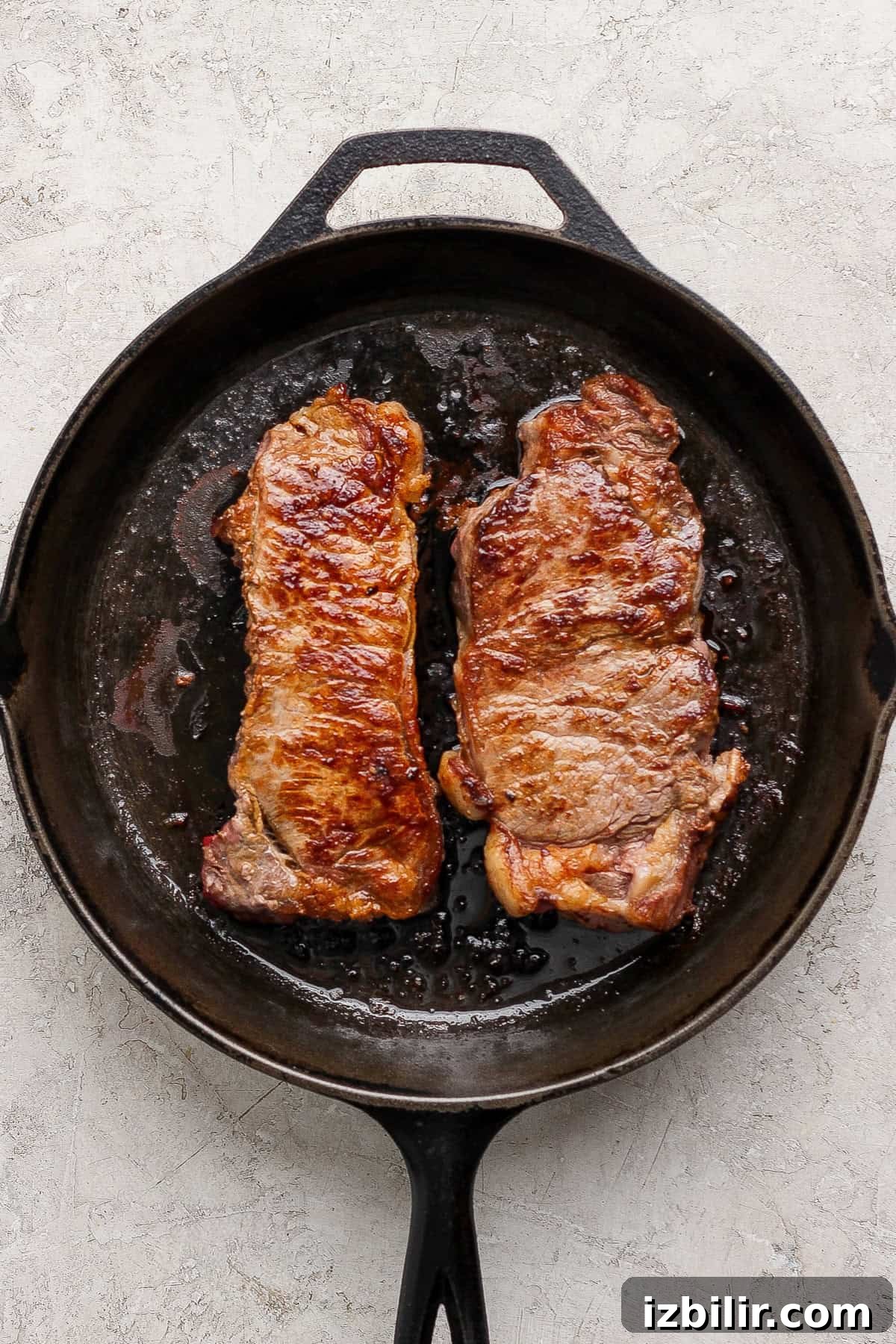 Two seared steaks are shown cooking in a black cast iron skillet on a light textured surface.