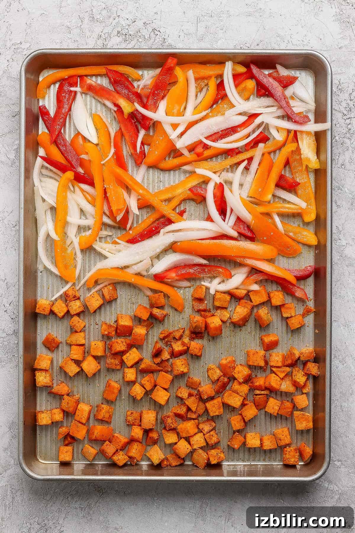 Sliced onions and bell peppers with seasoned diced sweet potatoes arranged on a baking sheet, ready for roasting.