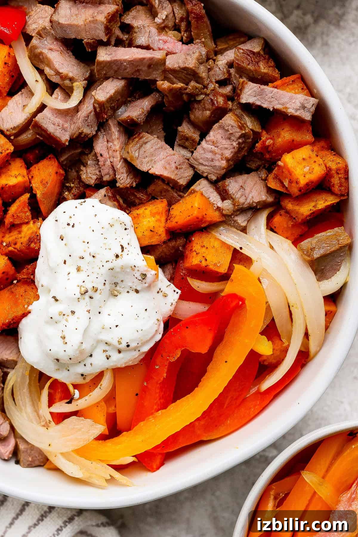 A bowl containing sliced steak, roasted sweet potatoes, sautéed onions, red and yellow bell peppers, and a dollop of sour cream, topped with cracked black pepper.