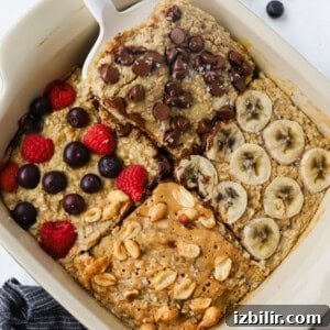A dish of baked oatmeal is adorned with four tempting toppings: chocolate chips, banana slices, berries, and peanuts drizzled with peanut butter. A serving is being carefully lifted with a spatula, showcasing its delicious layers.