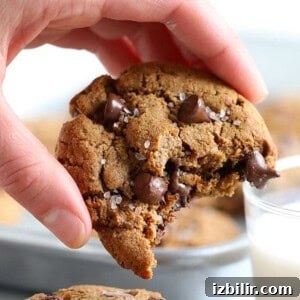 Wholesome Chocolate Chip Cookie Recipe 2 A hand holding a paleo chocolate chip cookie with a bite taken out, next to a glass of milk.