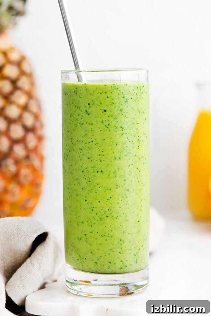 Tropical Spinach Smoothie with Pineapple and Mango 2 A spinach smoothie in a glass next to oranges and pineapples.
