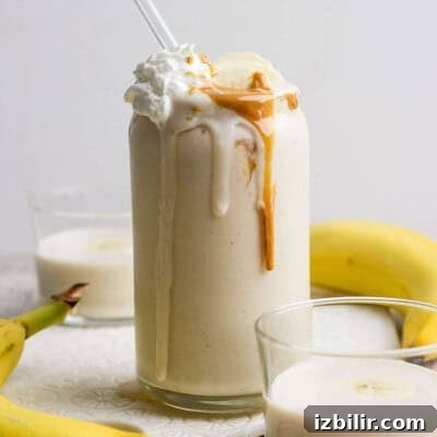 A glass of peanut butter banana protein shake topped with whipped cream, banana slices, and peanut butter, with a straw, surrounded by bananas and two smaller glasses of smoothie.
