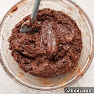 A glass bowl filled with thick, chocolate cookie dough and a gray spatula resting inside.