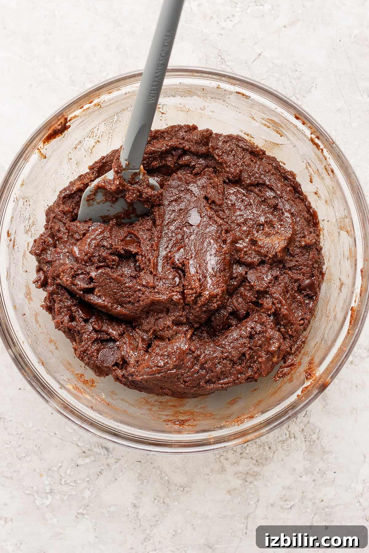 A glass bowl filled with thick, chocolate cookie dough and a gray spatula resting inside.