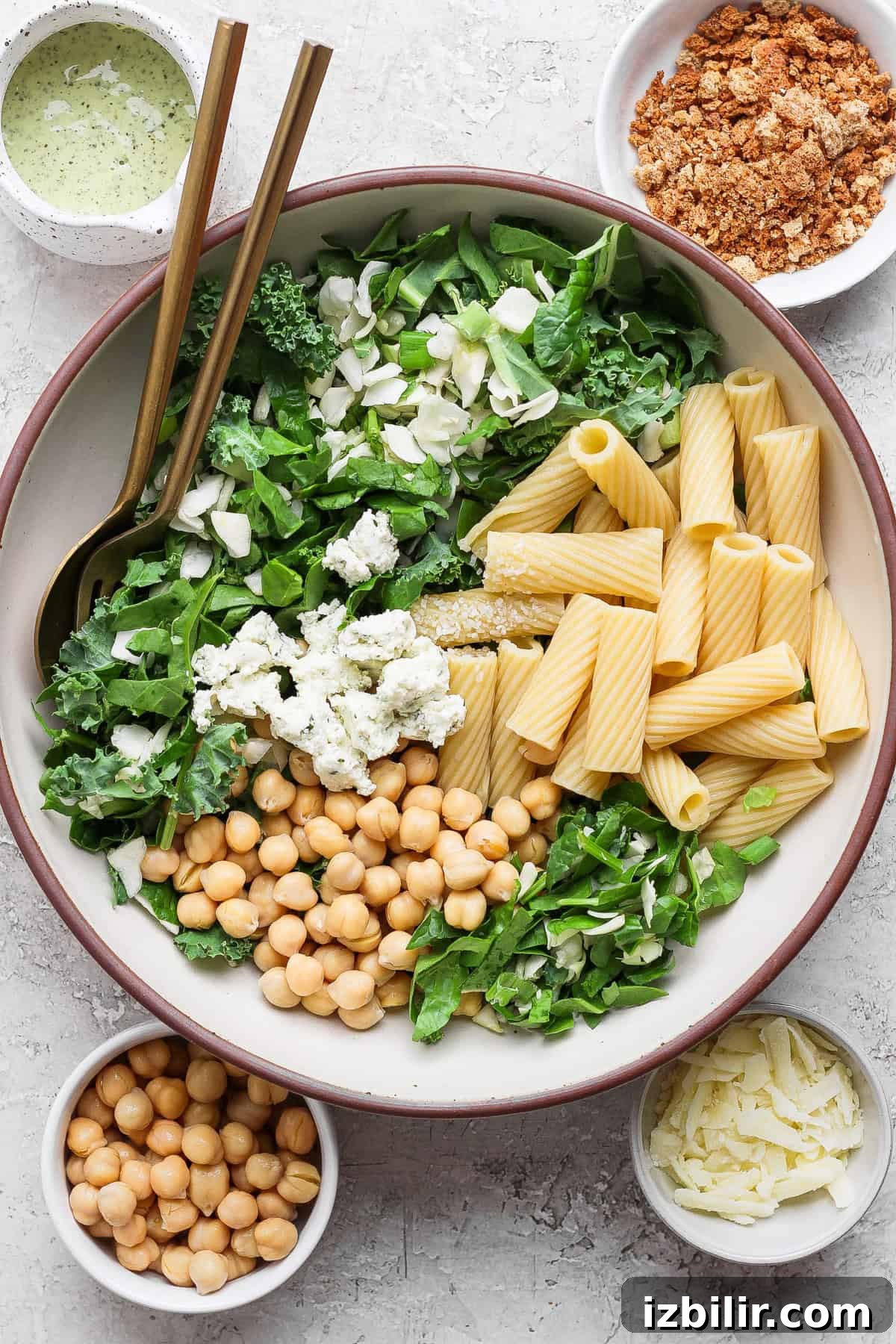 Green Goddess Pasta Salad: 6 Ingredients, No Dressing Needed 3 A large bowl with rigatoni pasta, chickpeas, leafy greens, feta cheese, and shaved parmesan, surrounded by bowls of dressing, crumbled topping, extra chickpeas, and parmesan.