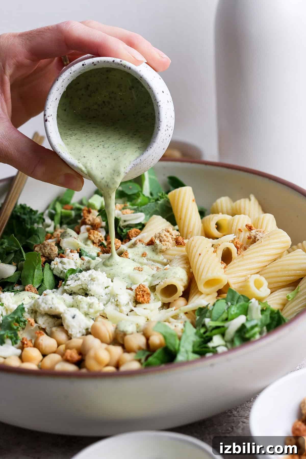 Green Goddess Pasta Salad: 6 Ingredients, No Dressing Needed 4 A hand pours creamy green dressing over a bowl of pasta salad with rigatoni, chickpeas, leafy greens, and crumbled toppings.