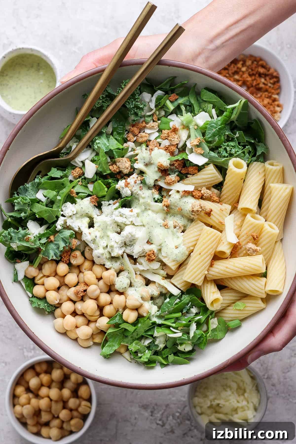 Green Goddess Pasta Salad: 6 Ingredients, No Dressing Needed 5 A bowl containing mixed greens, rigatoni pasta, chickpeas, cheese, and a creamy dressing, with two forks. Surrounding bowls hold extra chickpeas, shredded cheese, dressing, and crunchy toppings.