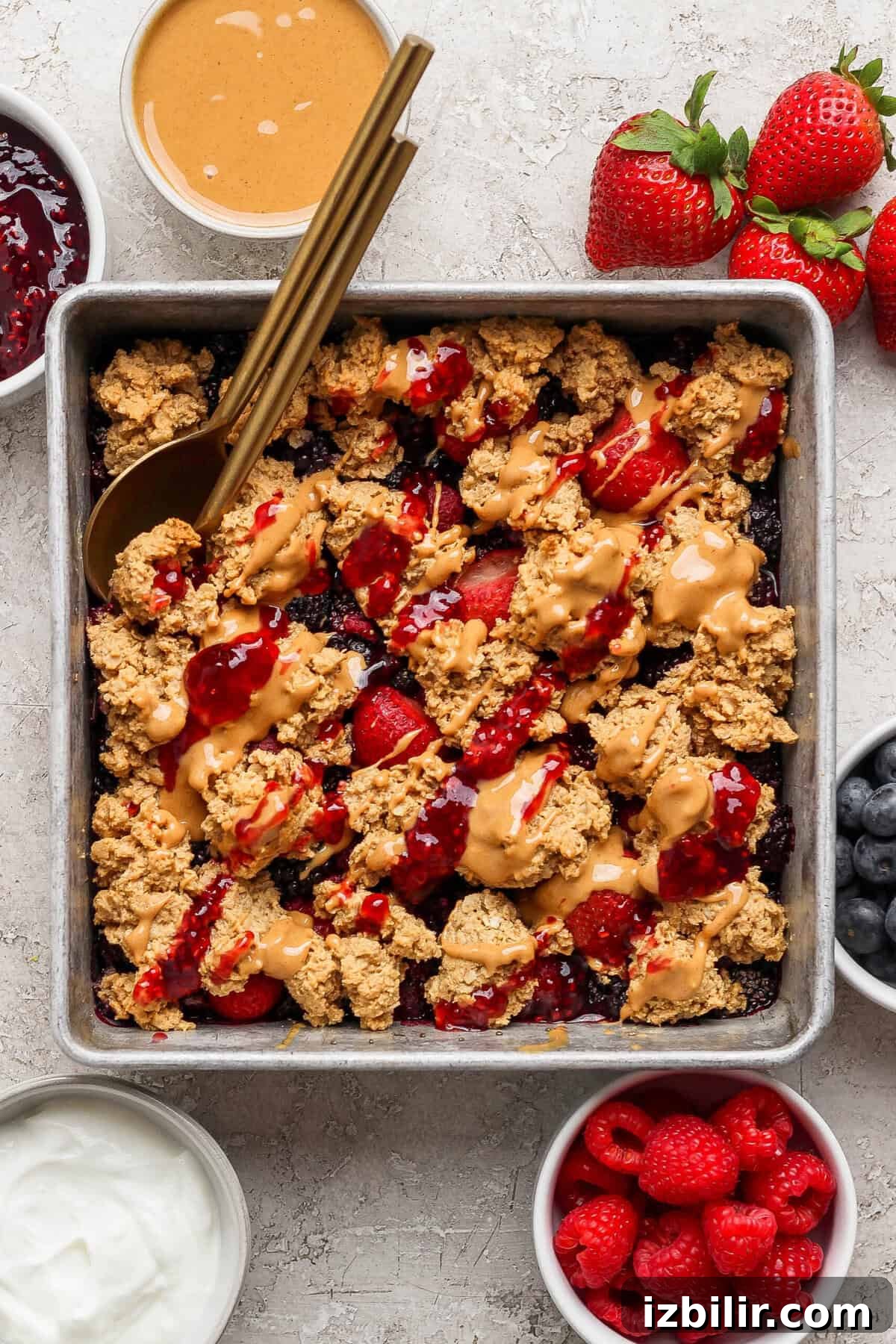 Peanut Butter & Jelly Protein Breakfast Crumble 7 overhead view of protein berry crumble in a pan with peanut butter drizzle surrounded by bowls of jam yogurt and fresh berries