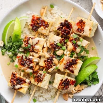A dish with skewered tofu topped with chili sauce on rice, garnished with lime wedges, green onions, peanuts, and herbs.