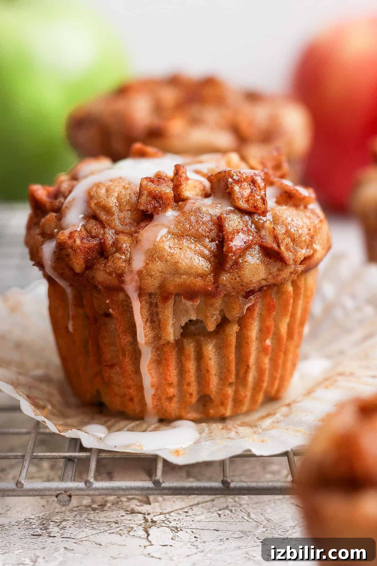 Protein-Packed Apple Muffins with Cottage Cheese 2 Close-up of an apple cinnamon muffin with icing on top, unwrapped and resting on a cooling rack, with apples blurred in the background.