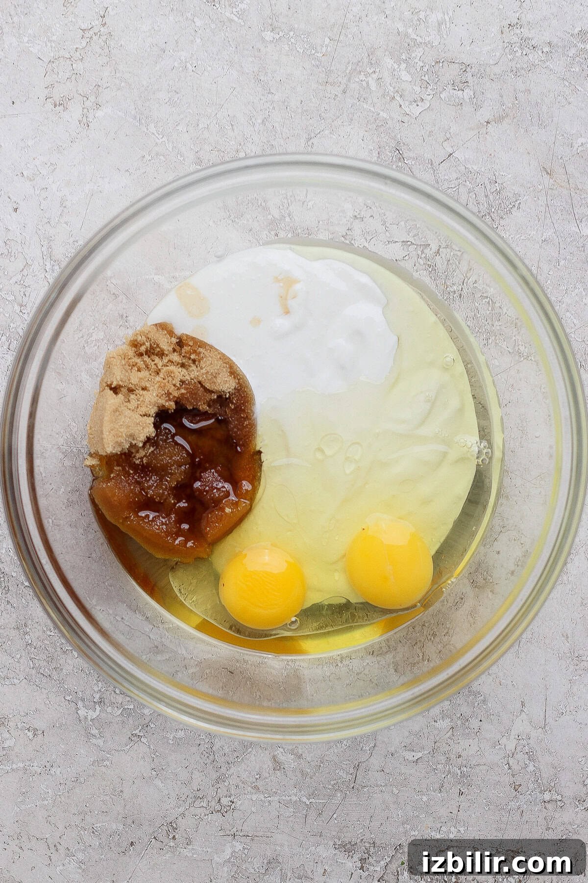 Protein-Packed Apple Muffins with Cottage Cheese 4 A glass bowl containing two eggs, brown sugar, honey, and a white liquid ingredient on a light textured surface.