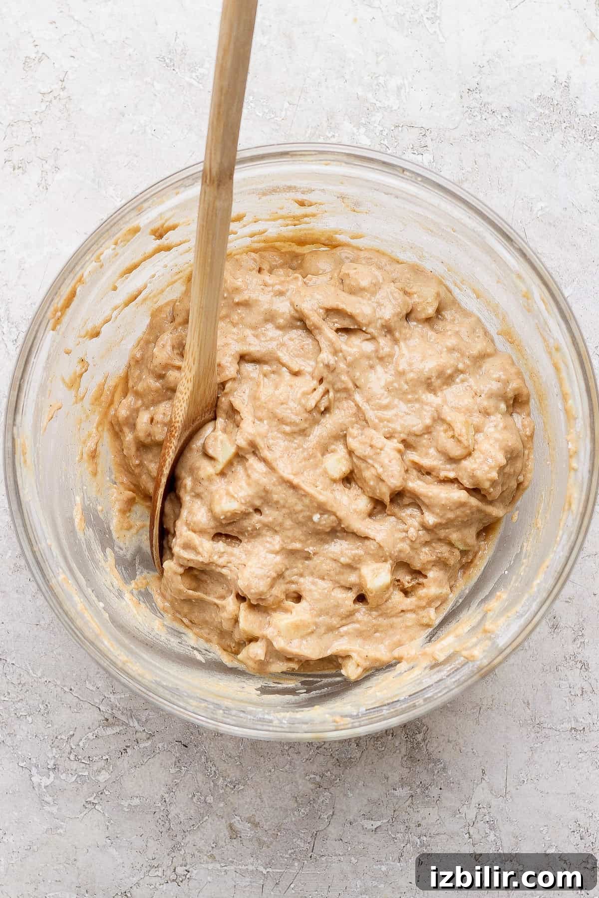 Protein-Packed Apple Muffins with Cottage Cheese 5 A glass bowl containing thick, chunky batter and a wooden spoon, set on a light-textured surface.