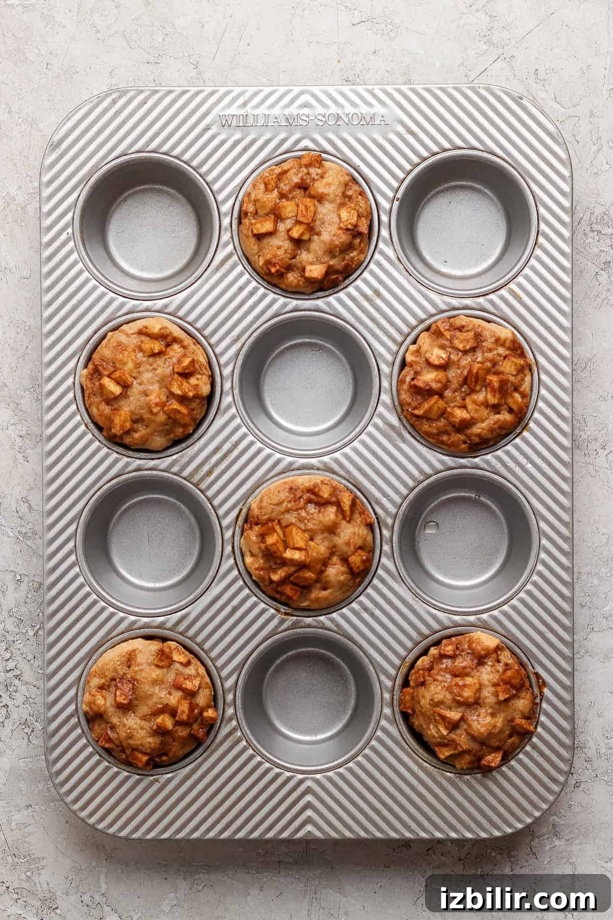 Protein-Packed Apple Muffins with Cottage Cheese 6 A muffin tin containing six baked muffins with diced apple pieces, with three empty spaces in the tray, on a light textured surface.