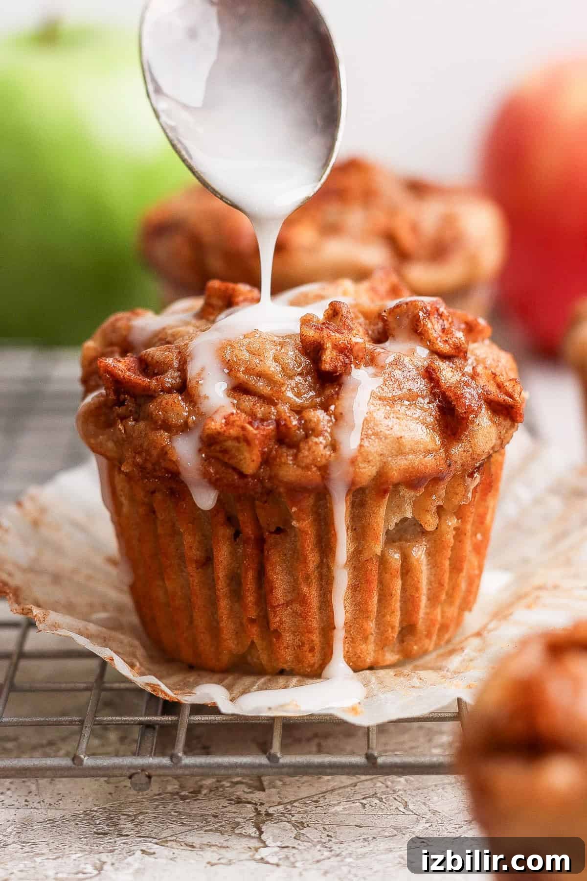 Protein-Packed Apple Muffins with Cottage Cheese 7 A spoon drizzles white icing over a muffin topped with small pieces of fruit, set on a wire rack with an open paper liner.