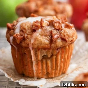 A muffin topped with small cinnamon apple pieces and white icing, sitting on an opened paper liner with a blurred background.
