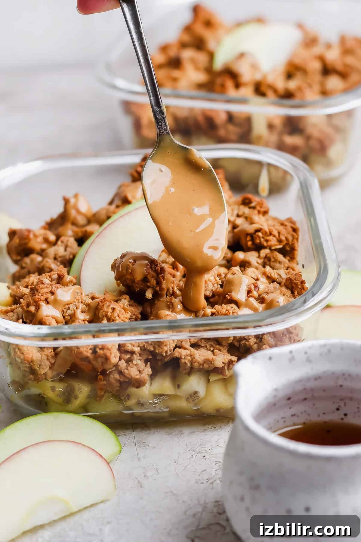Nut butter drizzled over protein apple crisp in an individual glass meal prep container with apple slices.