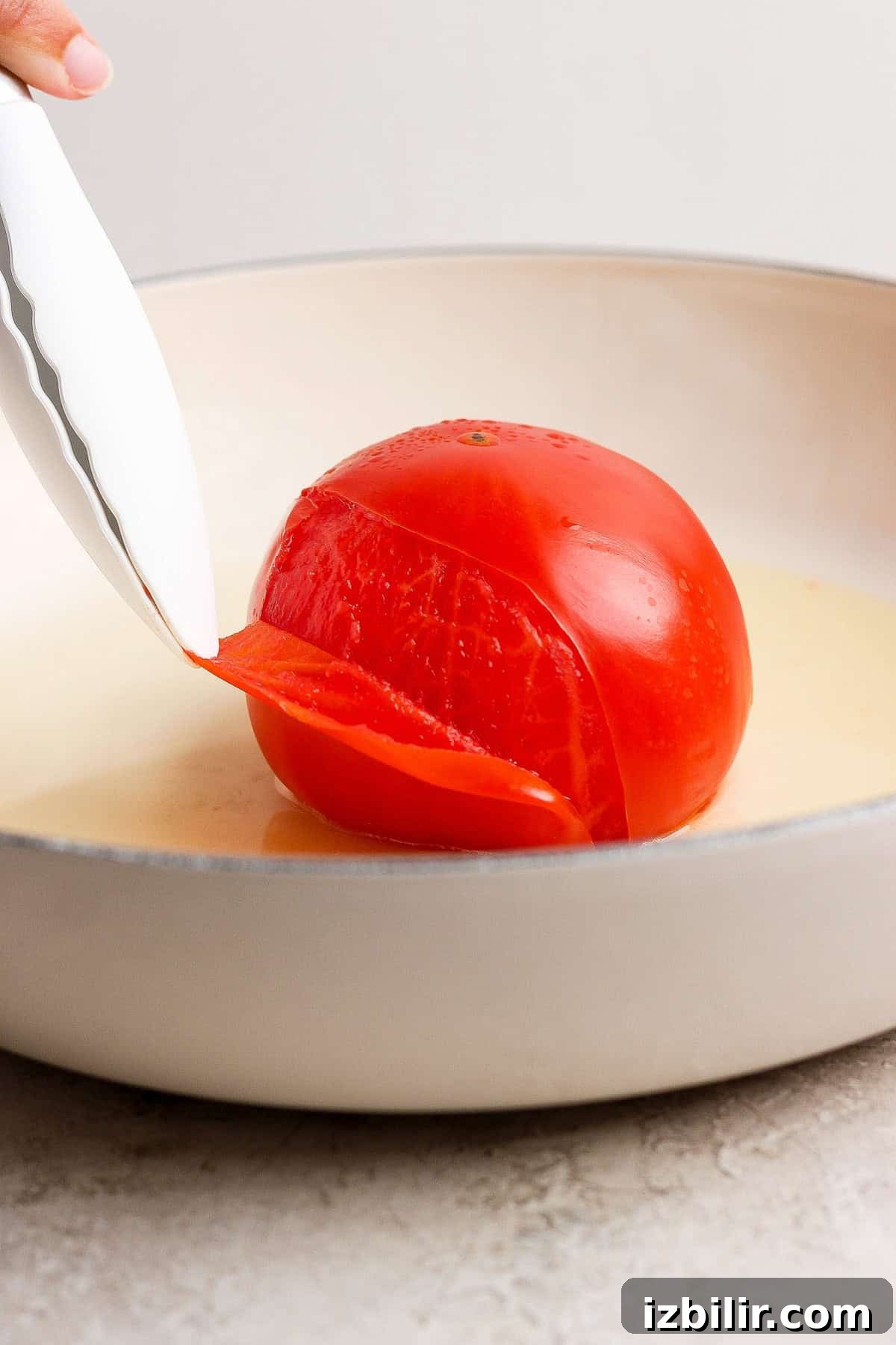 Tongs removing the loosened peel from a steamed whole tomato in a skillet.