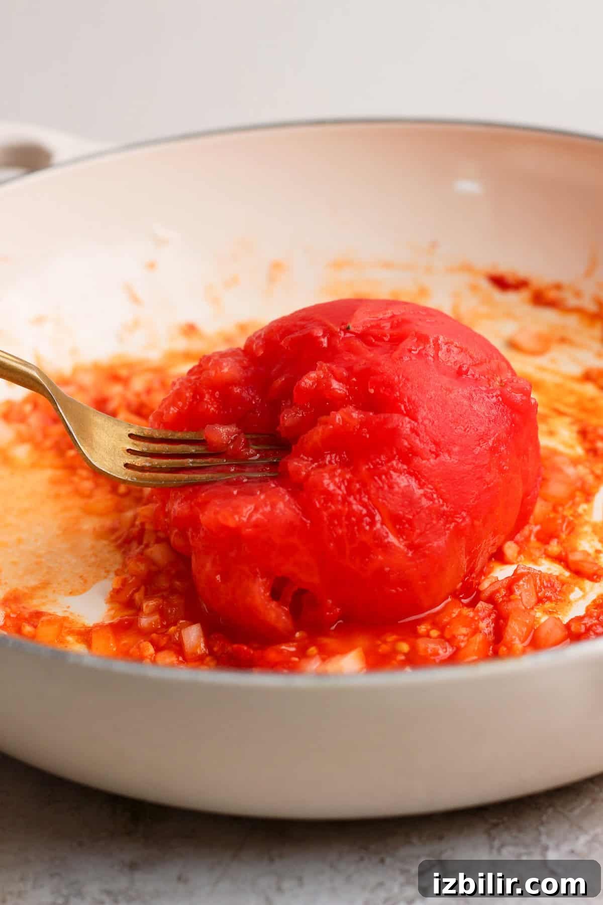 A peeled steamed tomato being smashed into onion, garlic, and tomato paste with a fork.