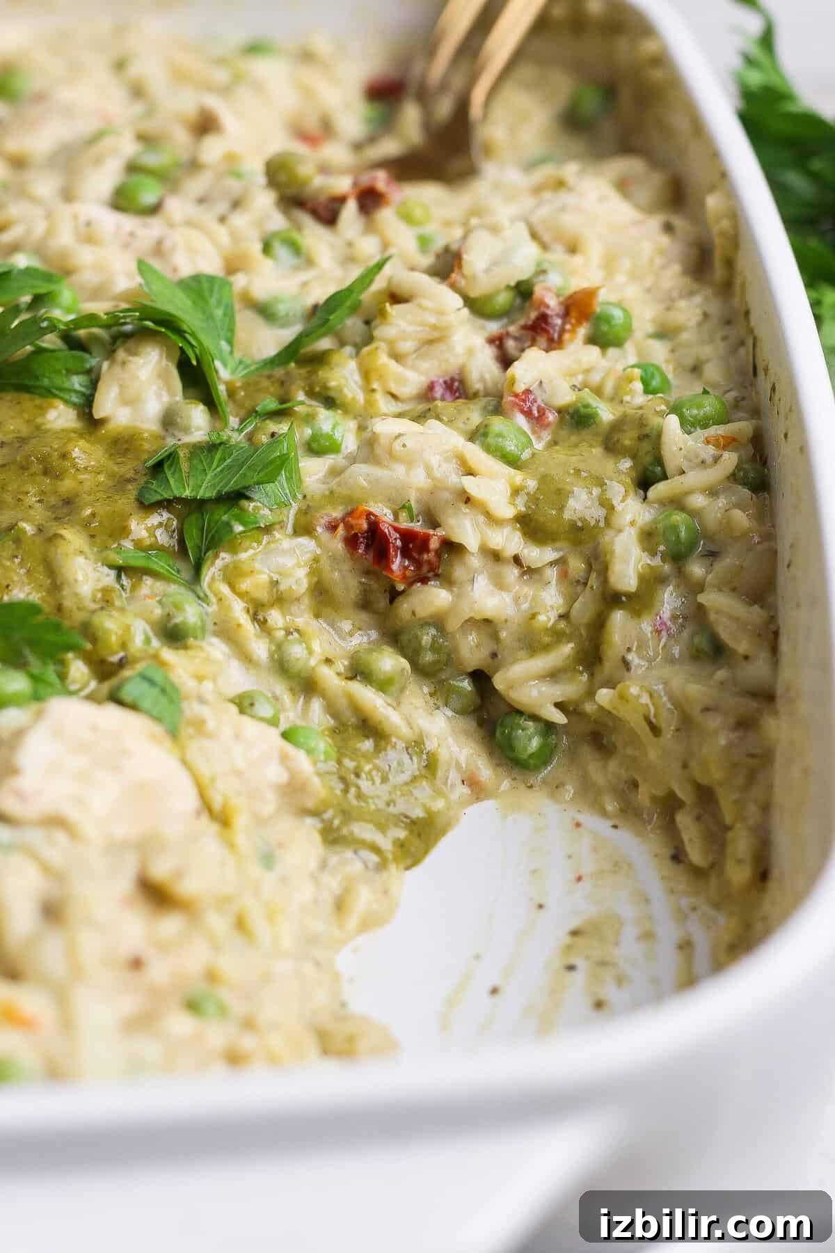 Dump-and-Bake Pesto Chicken Orzo 2 A serving spoon scooping creamy chicken pesto orzo with peas, sun-dried tomatoes, pesto, and fresh parsley from a white casserole dish.