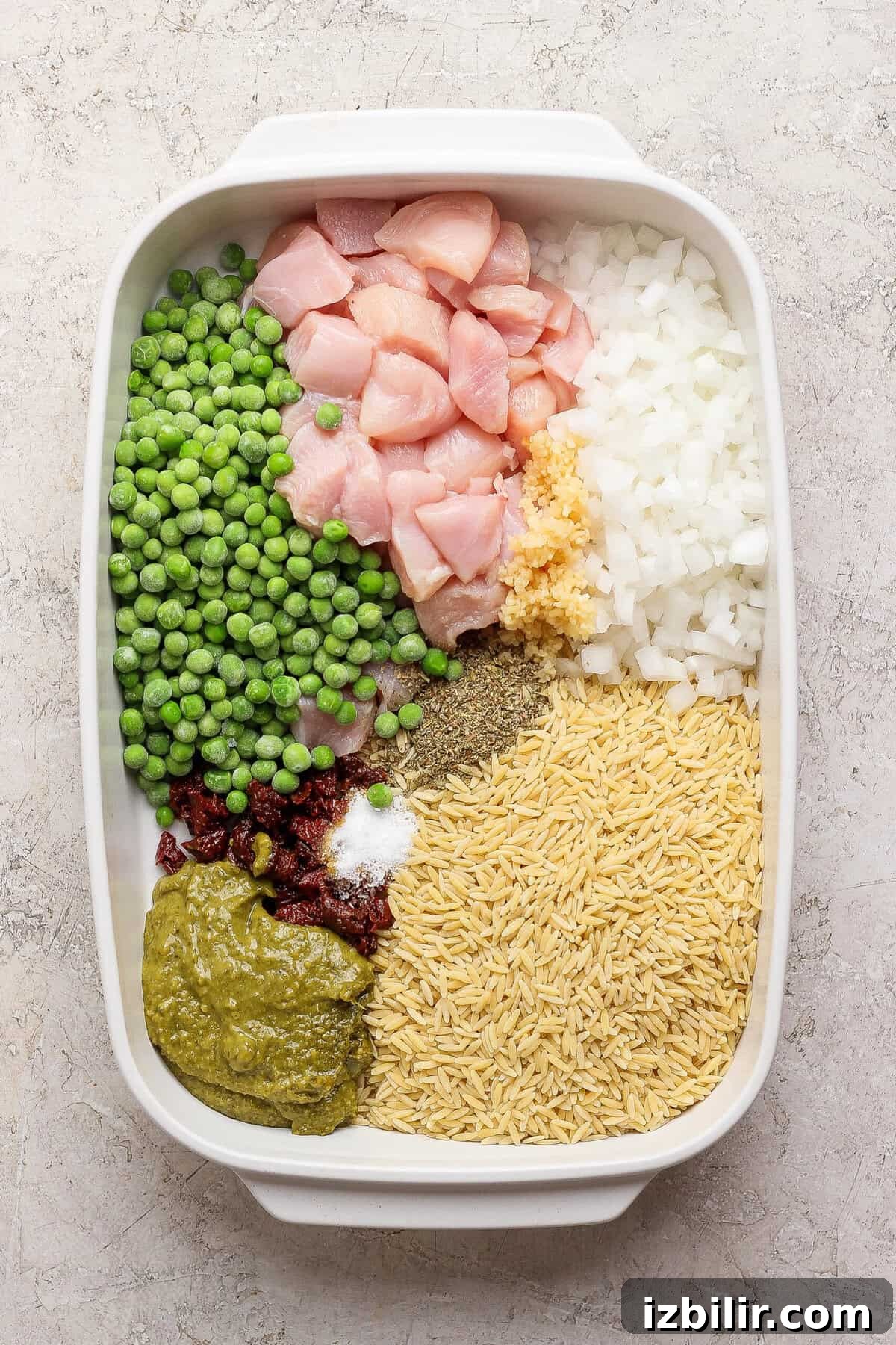 Dump-and-Bake Pesto Chicken Orzo 3 Raw chicken, dry orzo, frozen peas, onion, garlic, pesto, sun-dried tomatoes, and seasonings arranged in a white casserole dish.