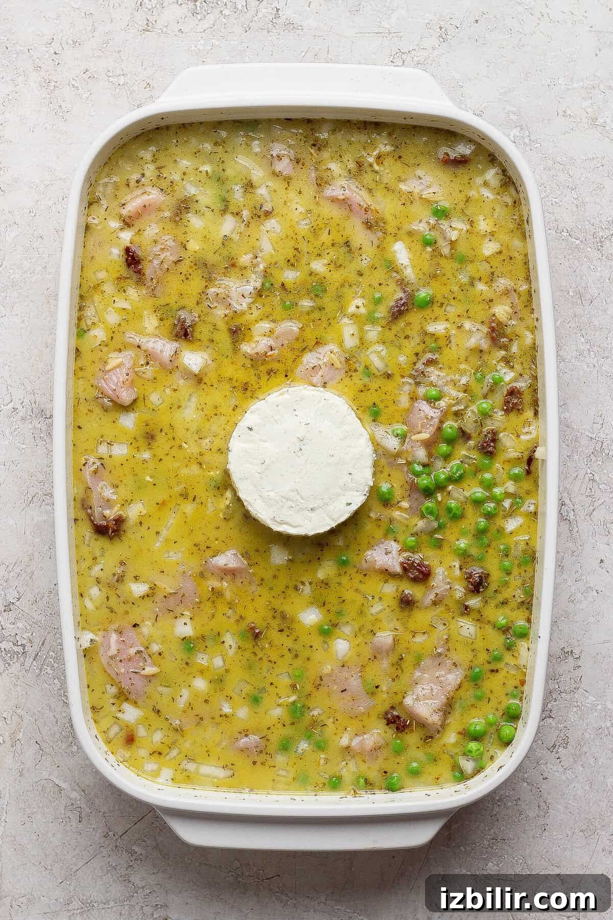 Dump-and-Bake Pesto Chicken Orzo 5 Unbaked chicken pesto orzo mixture in a white casserole dish with a whole puck of Boursin in the center.