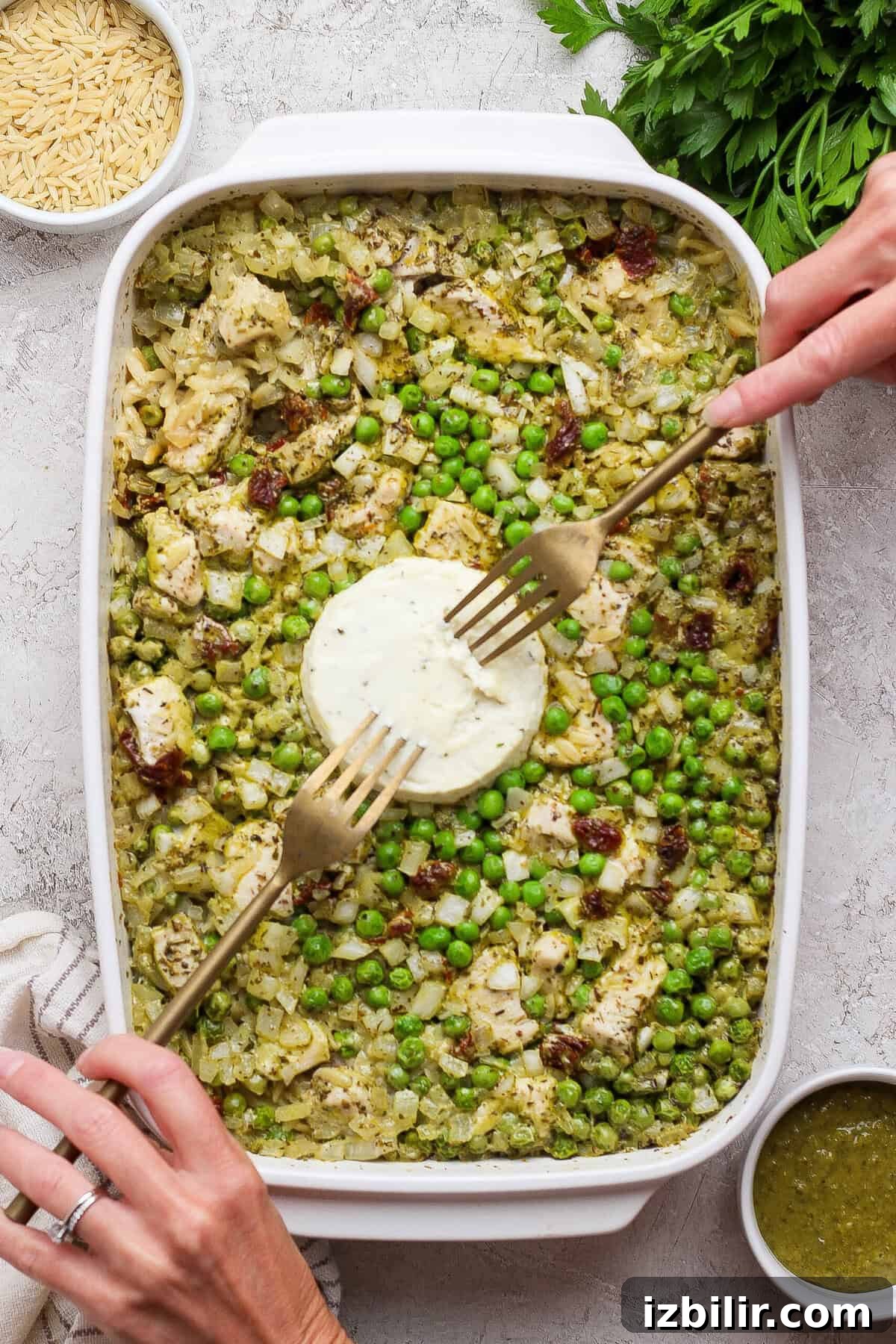 Dump-and-Bake Pesto Chicken Orzo 6 Hands using forks to mix softened Boursin into baked chicken pesto orzo with peas and sun-dried tomatoes.