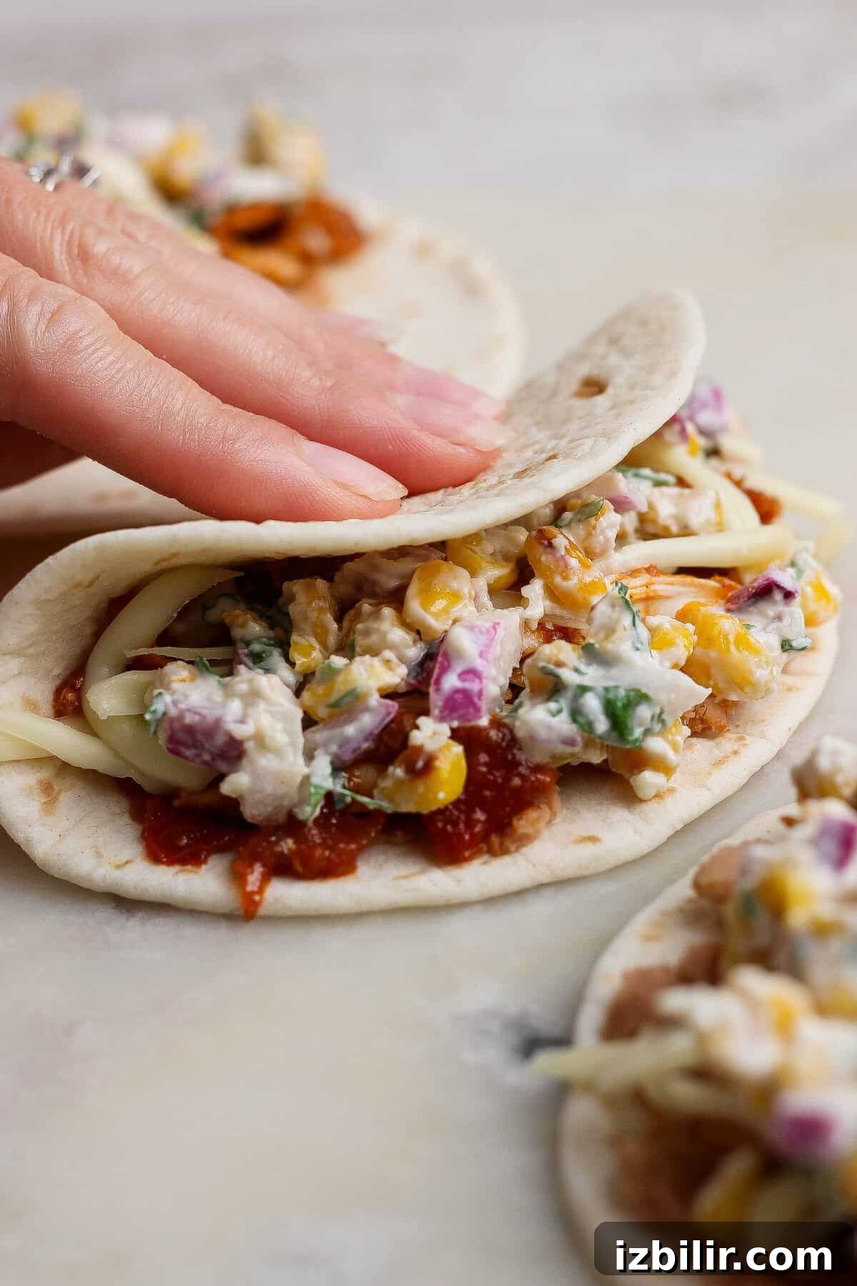 Oven-Baked Street Corn Chicken Tacos 5 A hand folding a filled flour tortilla over street corn salad, shredded chicken, refried beans, and cheese.