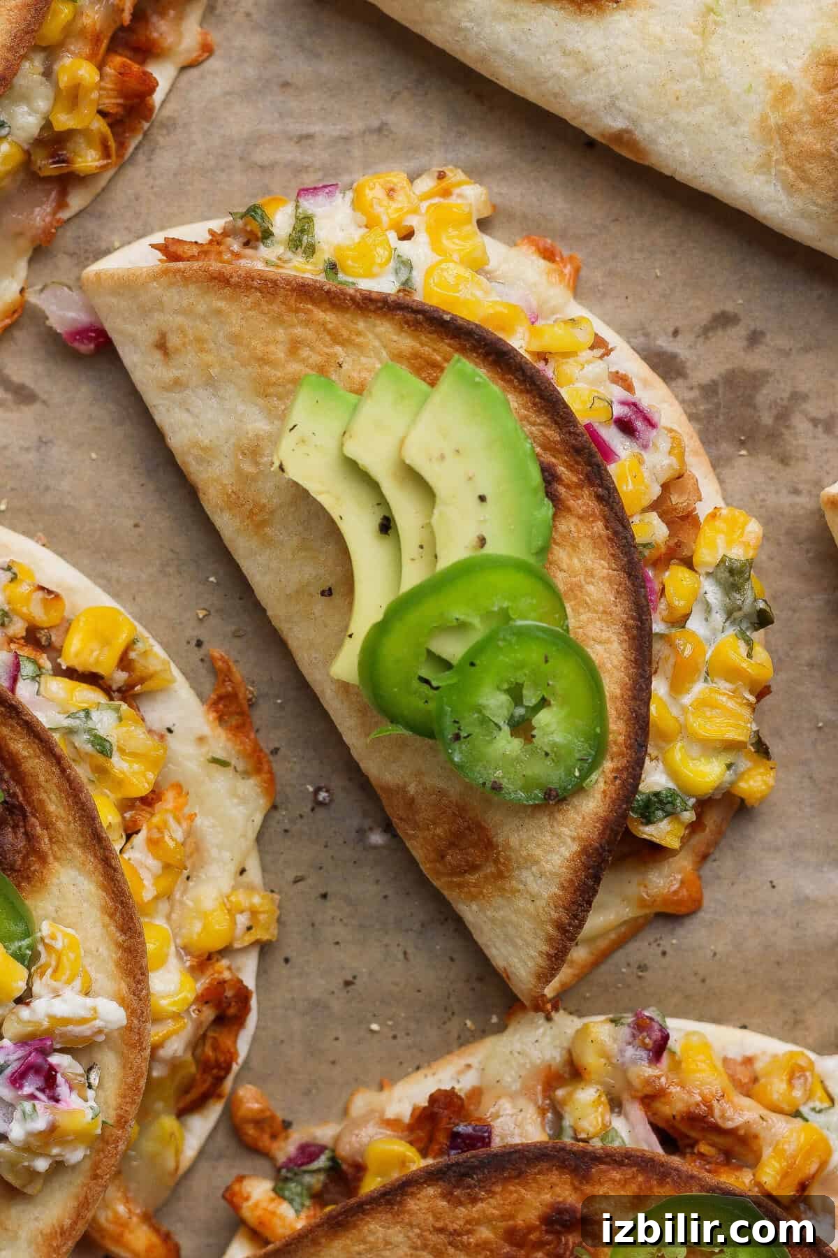 Oven-Baked Street Corn Chicken Tacos 7 Crispy street corn chicken tacos topped with sliced avocado and jalapeño on a parchment-lined baking sheet.