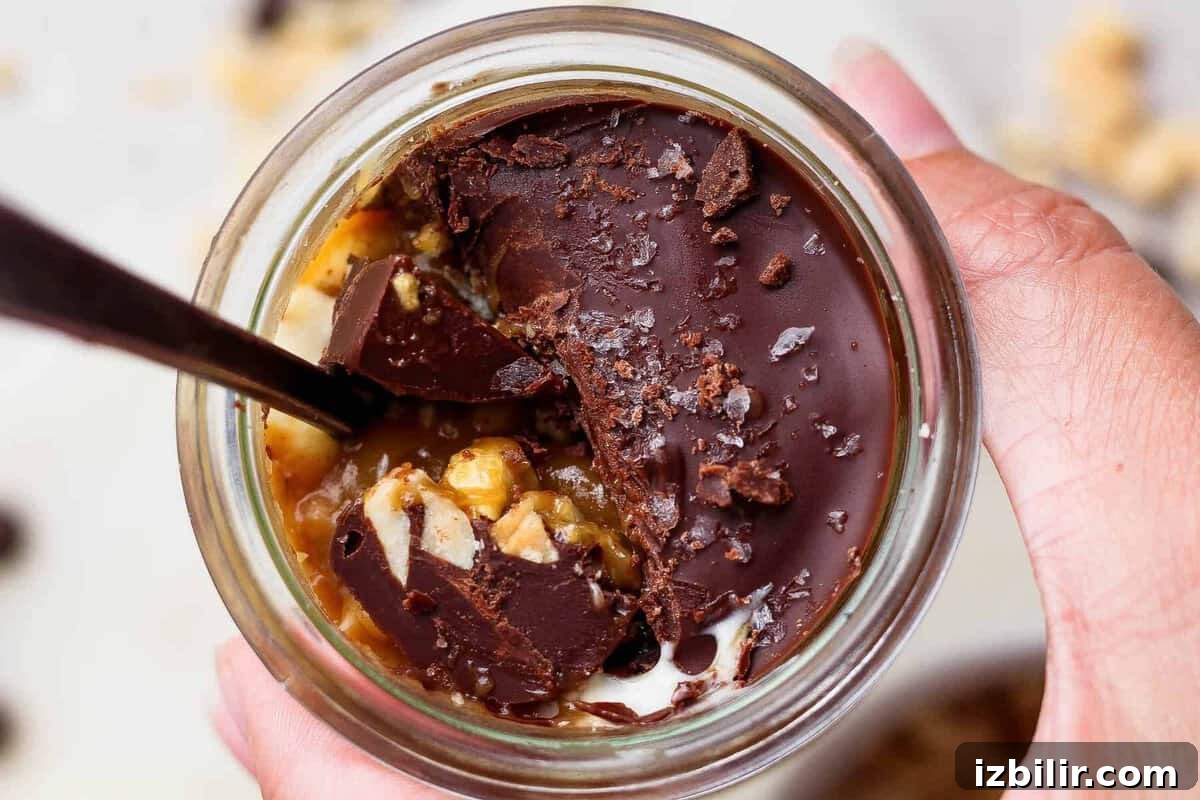 High-Protein Snickers Cheesecake Cups Recipe 2 Spoon breaking through the chocolate shell on an individual Snickers cheesecake cup to reveal peanut butter and cottage cheese layers.