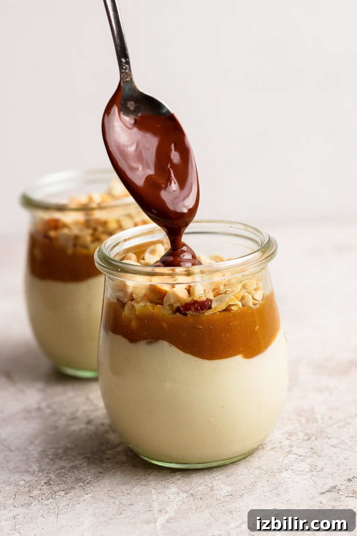 High-Protein Snickers Cheesecake Cups Recipe 7 Melted chocolate magic shell being spooned over peanut butter and chopped peanuts in individual cheesecake cups.