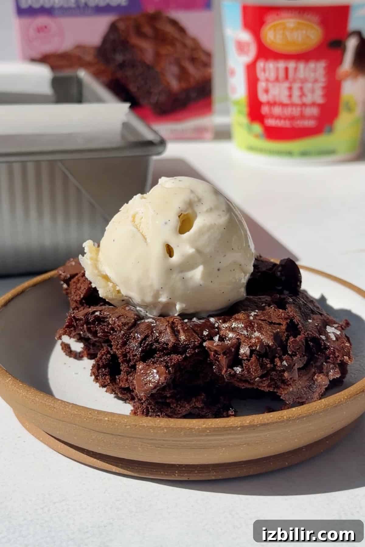 Fudgy 2-Ingredient Cottage Cheese Brownies 5 Cottage cheese brownie topped with vanilla ice cream, with the brownie pan and a cottage cheese container in the background.