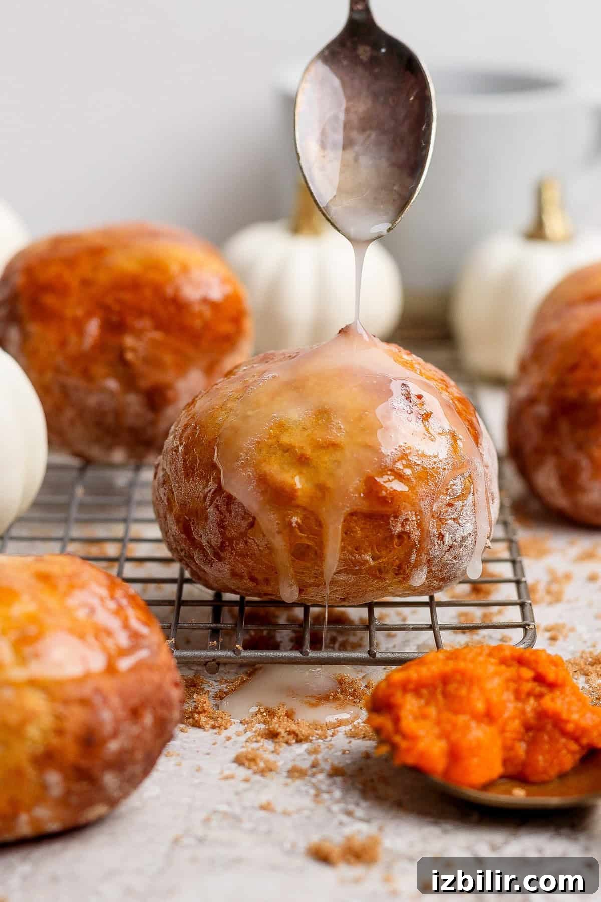 Crispy Cottage Cheese Pumpkin Fritters Recipe 10 Spoon drizzling white glaze over a round pumpkin fritter on a cooling rack with more fritters and pumpkins behind it.