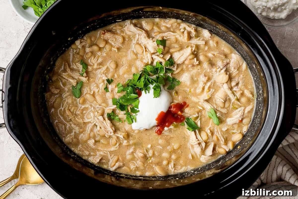 Slow cooker filled with white chicken chili topped with cottage cheese, hot sauce, and chopped fresh herbs.