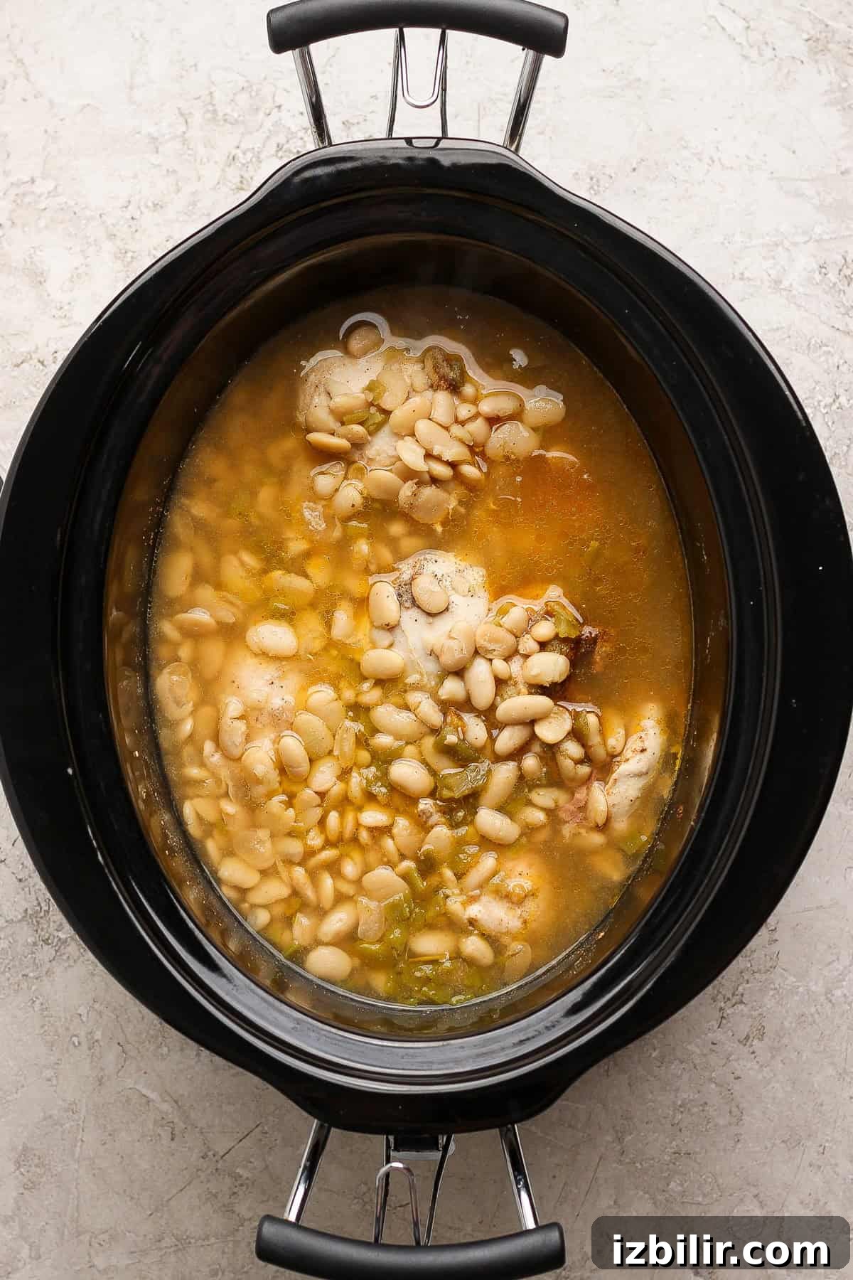 Cooked chicken and white beans in their cooking liquid inside a slow cooker before shredding.