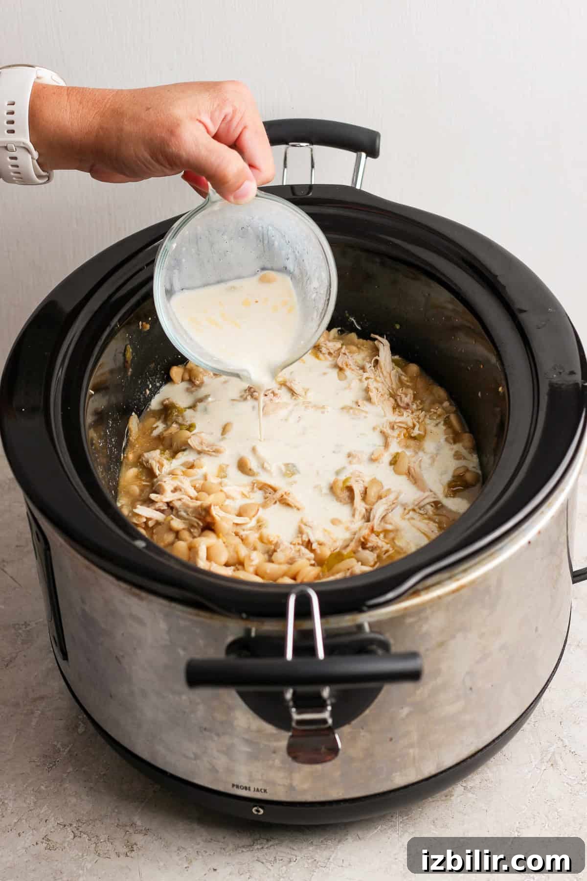 Creamy cottage cheese mixture being poured into a slow cooker of shredded chicken and white beans.